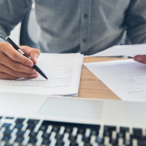 Unrecognizable businessman sitting at table holding ballpoint pen and papers and checking documents in office