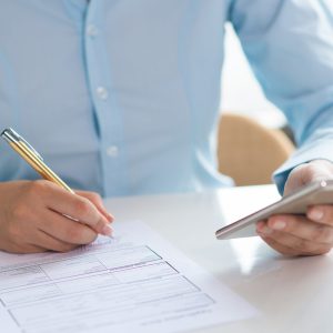 Closeup of woman signing document and holding smartphone. Document lying on table. Contract or application concept. Cropped view.